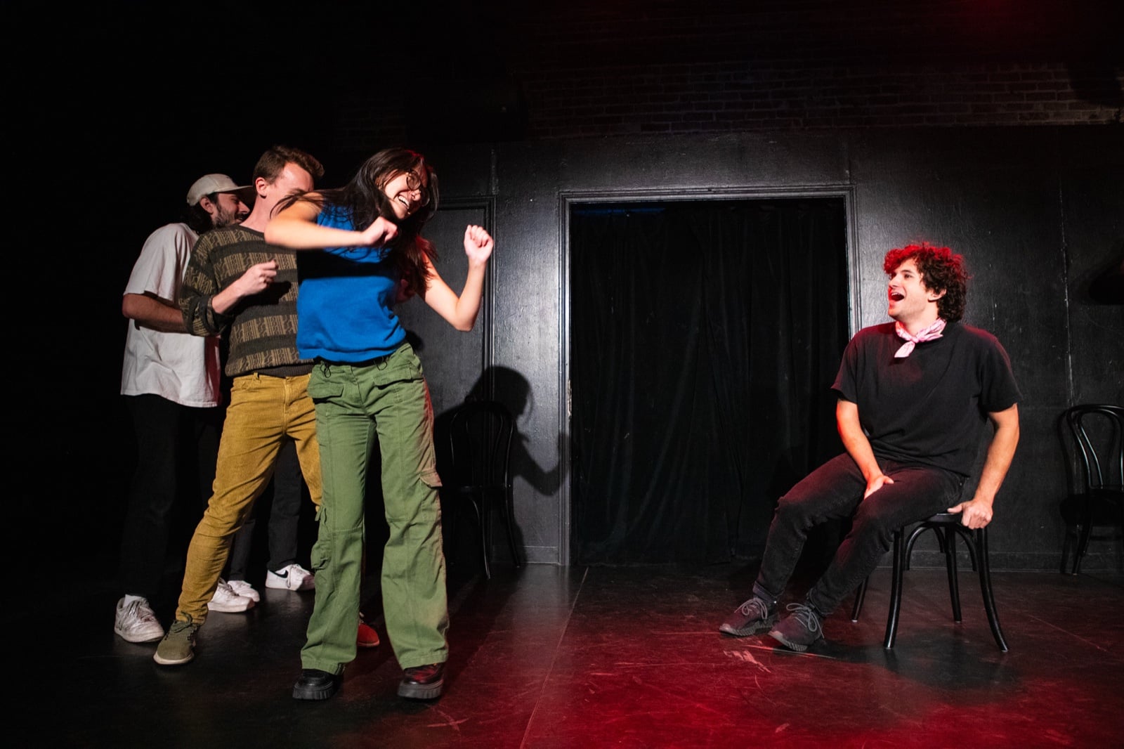 Improv performers mid-scene on a dark theater stage with dramatic lighting.