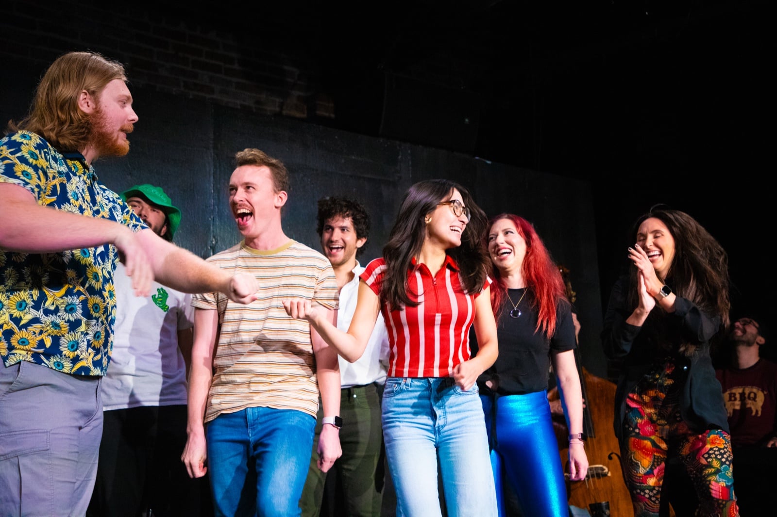 Ensemble improv cast laughing together on stage under spotlights.